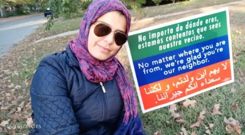 A photo of one of our students next to a sign with text in three languages, Spanish, English and Arabic. The English part says "No matter where you are from, we're glad you're our neighbor"