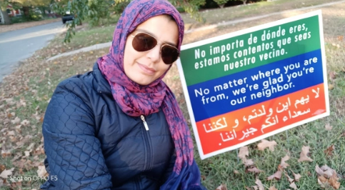 A photo of one of our students next to a sign with text in three languages, Spanish, English and Arabic. The English part says "No matter where you are from, we're glad you're our neighbor".