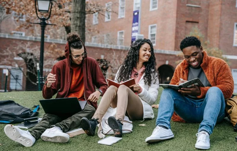 Three students sitting on the lawn on a college campus studying and talking.