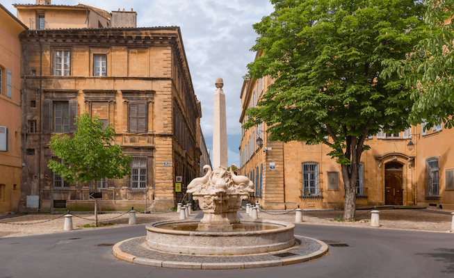 An ornate round marble fountain with fish spouting water in the middle of a square in a quaint French town.