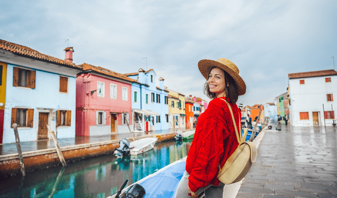 A woman in a red sweater and straw hat standing next to a canal in Italy.
