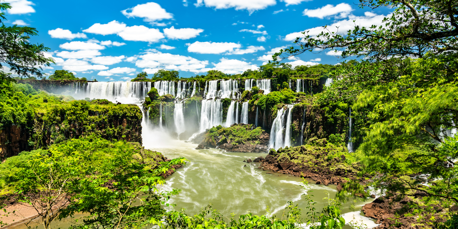 A wide and verdant view of Iguazu Falls in Argentina with blue skies and puffy clouds.