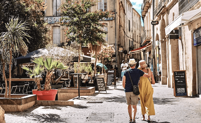 A man and woman in summer clothes walking arm in arm along an ancient cobble stone street with palm trees, shops and cafe's.