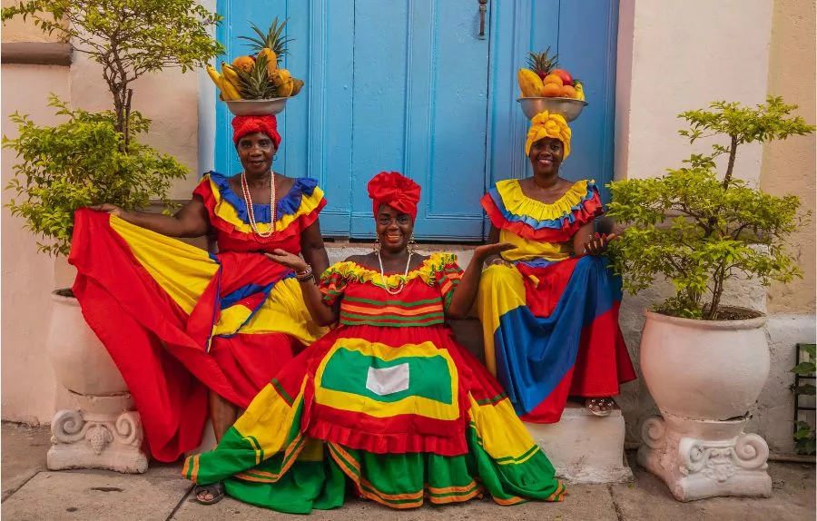 3 Colombian women wearing traditional dresses sitting on the stairs in front of a blue door