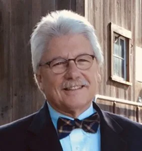 A man with white hair in a bow tie and suit wearing glasses