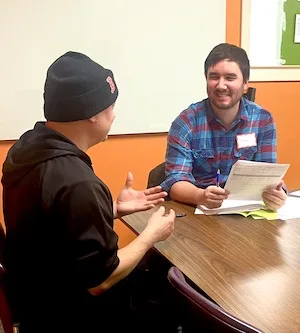 Two people sitting at a table in a classroom having a conversation and smiling.