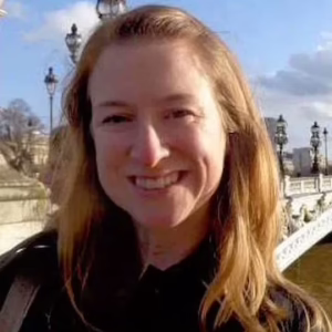 A smiling woman with light hair on a Parisian bridge.