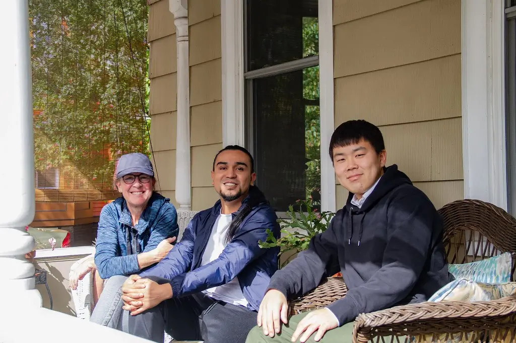 Two students sitting on a porch with a host smiling on a sunny day.