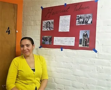 Student Blanca wearing a bright yellow top is standing next to a poster about Harriet Tubman on one of the white painted brick walls of her English language school, ILI in Massachusetts.