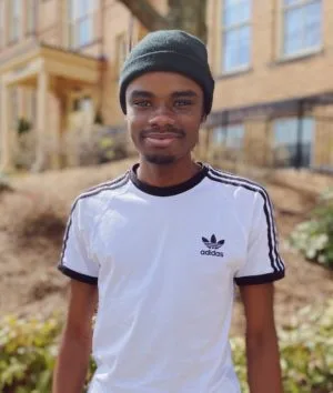 A photo of John Steve Papouto Felix, standing outside ILI's language school in Massachusetts. He's wearing a white adidas T-shirt with navy blue strips over the shoulders, and a beanie hat. He's looking straight into the camera lens and smiling.