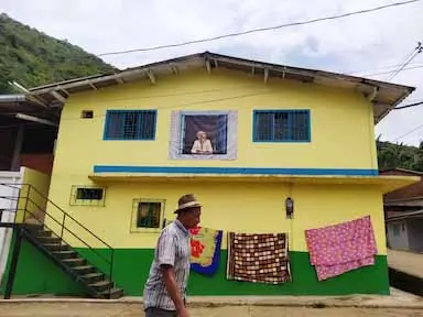 A man walking in front of a yellow house. There are blankets hanging to dry and a poster of a woman looking out a window on the wall.