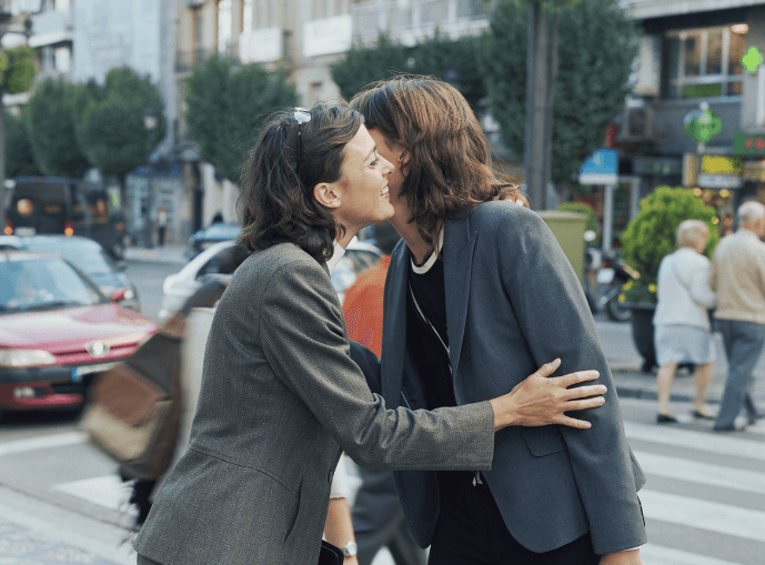 Two women greet each other with a kiss on the cheek on a lively street.