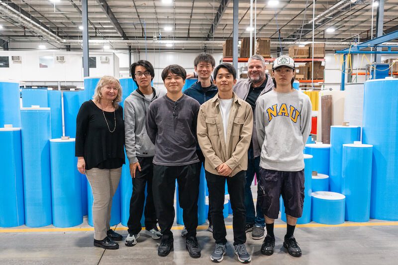 A group of people pose in front of blue rolls in a factory.