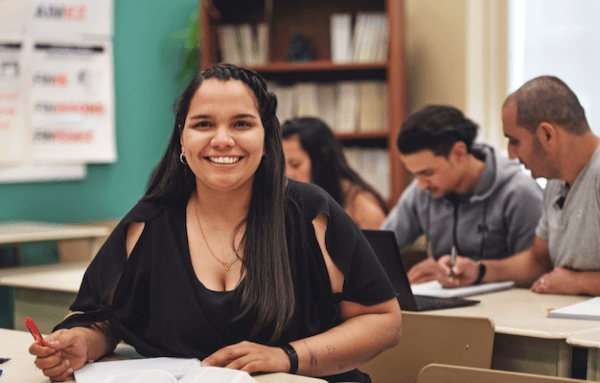 A woman smiles at her desk in a classroom, surrounded by students engaged in learning.