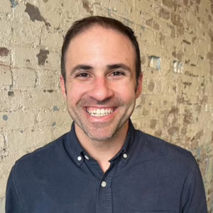 A smiling man dressed in a blue shirt looks directly at the camera, radiating positivity and friendliness.