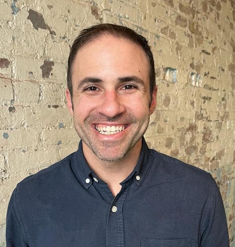 A smiling man dressed in a blue shirt looks directly at the camera, radiating positivity and friendliness.