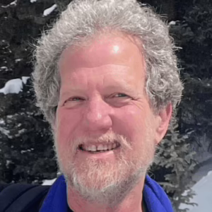 A man with a beard dressed in a blue shirt smiles for the camera, showcasing a friendly and inviting expression.