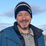 A man wearing a blue jacket and hat smiles warmly.
