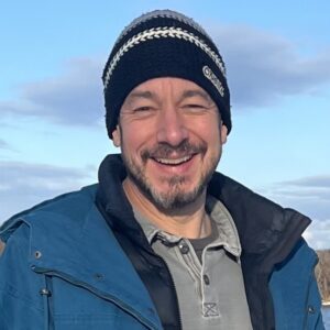 A man wearing a blue jacket and hat smiles warmly.