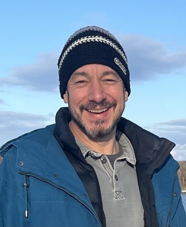 A man wearing a blue jacket and hat smiles warmly.