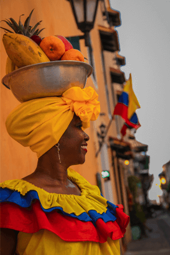 A woman wearing a vibrant dress balances a bowl of fruit on her head, showcasing a blend of color and culture.