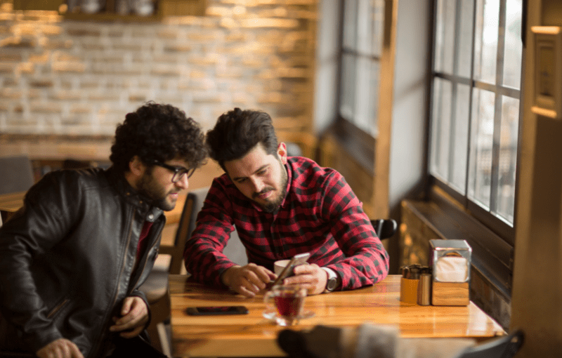 Two men seated at a wooden table in a cafe, looking at a smartphone.