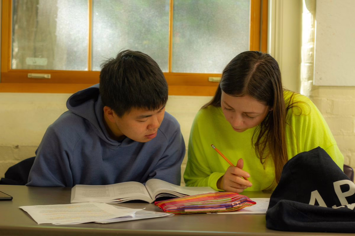 2 students sitting in a classroom doing an exercise on a book together.