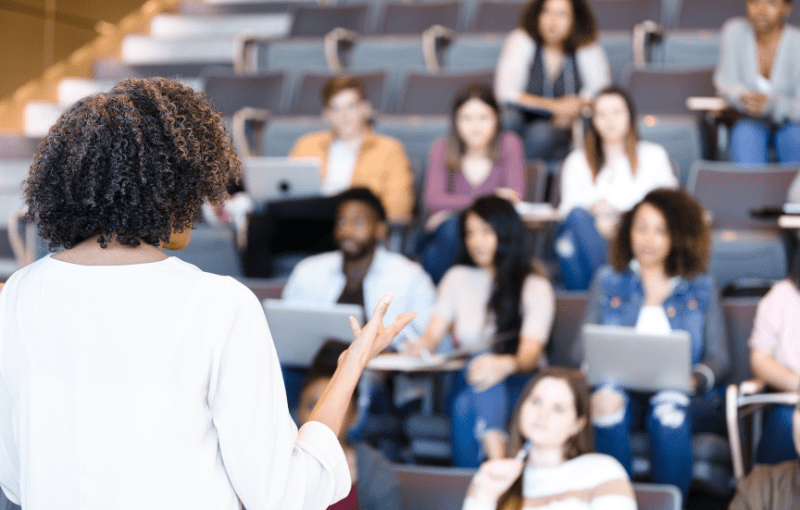 A woman speaks to a group of seated students in a lecture hall. Some students use laptops, while others listen attentively.