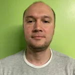 Portrait of a person in a grey shirt standing against a bright green wall, facing the camera.