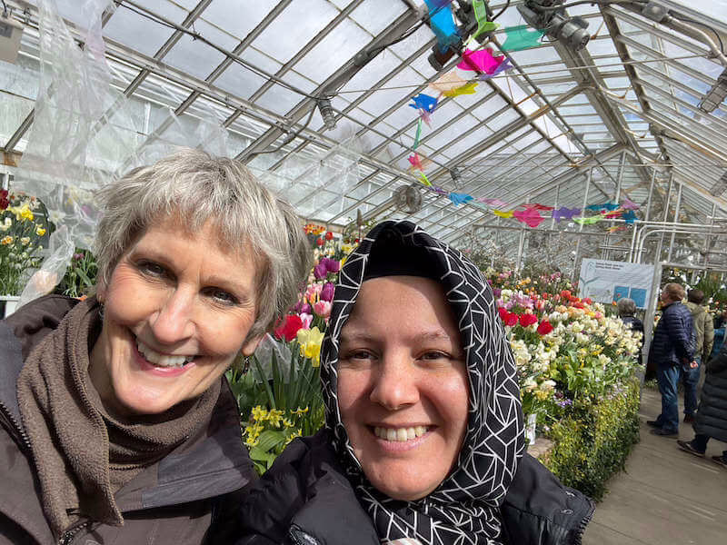 Two women smiling inside a greenhouse filled with colorful flowers.