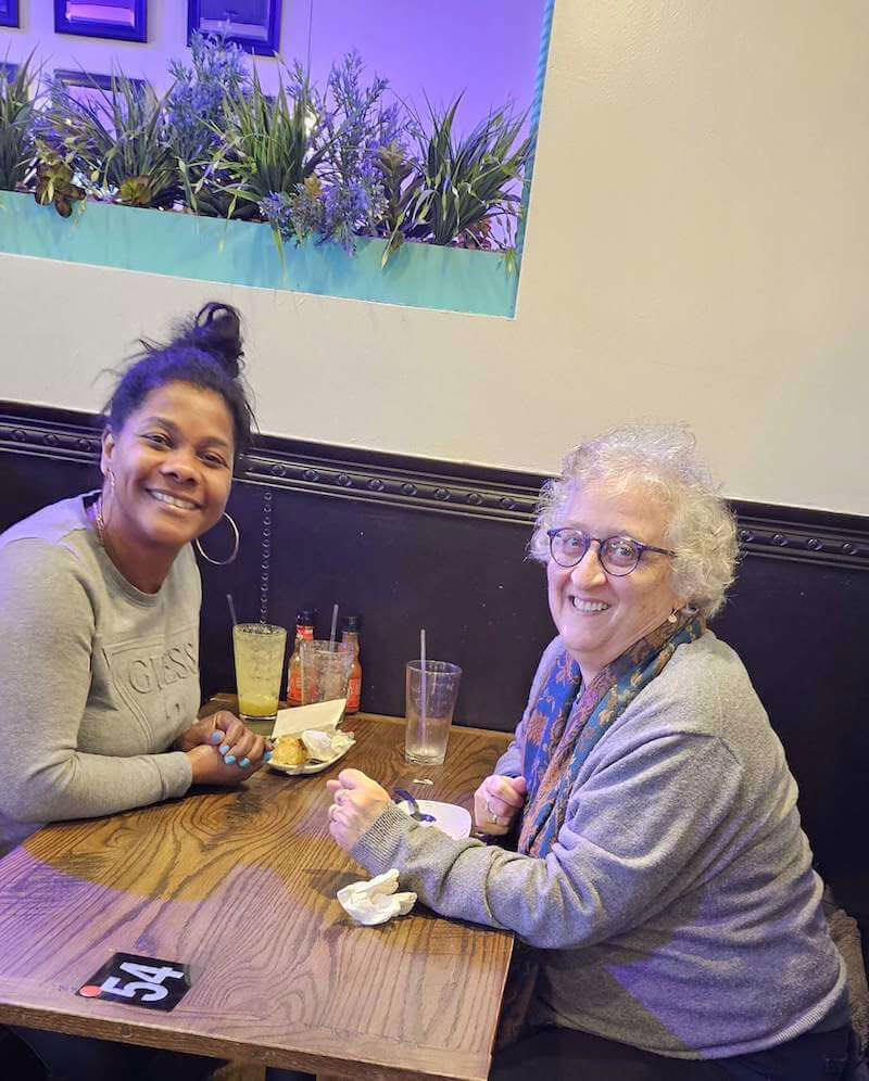 Two women sit at a table in a restaurant, smiling at the camera with drinks and food in front of them.