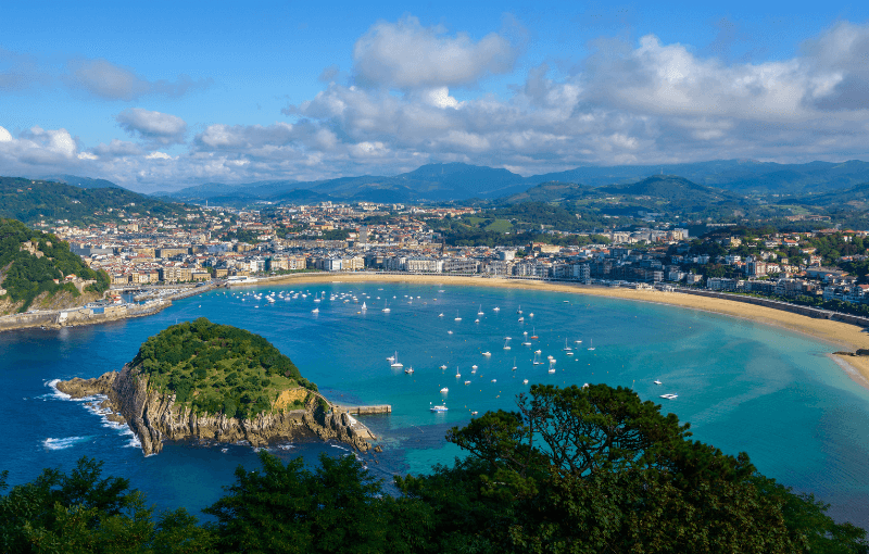A panoramic view of a coastal city with a sandy beach, turquoise bay, anchored boats, a small island, and mountains in the background under a partly cloudy sky.