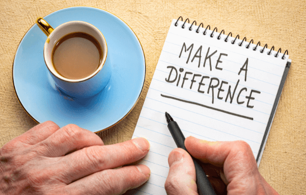 A person writes "MAKE A DIFFERENCE" in a notebook with a black marker, next to a cup of coffee on a table.