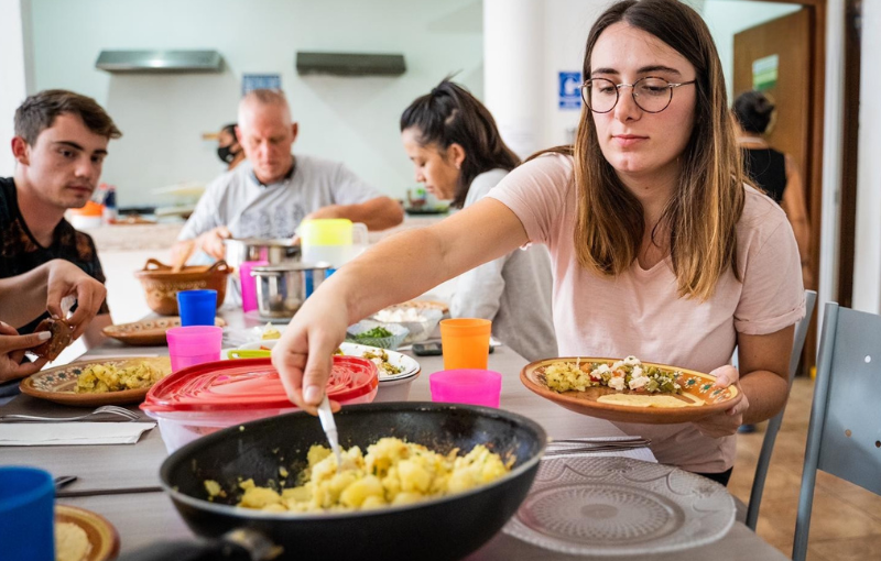 A woman serves herself some food at a table surrounded by other people engaged in conversation and cooking activities.