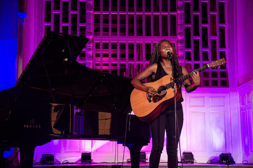 A woman on stage singing and playing an acoustic guitar in front of a piano.