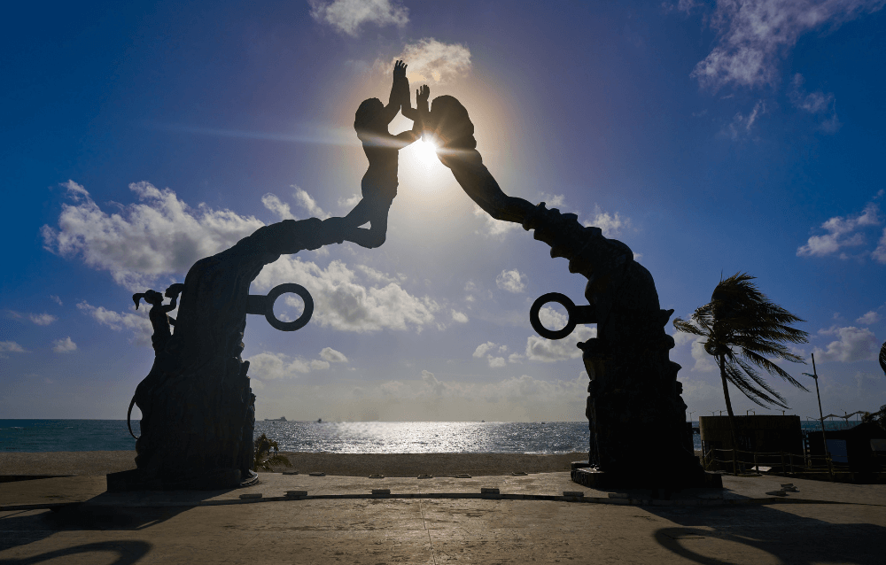 Two large statues of figures reaching toward each other form an archway by the ocean with the sun shining between their hands, silhouetted against a cloudy sky.