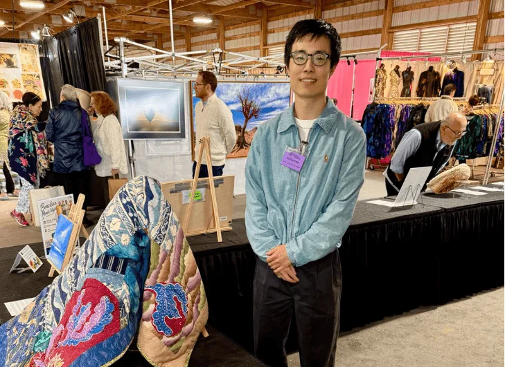 A man wearing glasses and a light blue jacket stands in front of a display booth at an arts and crafts fair.