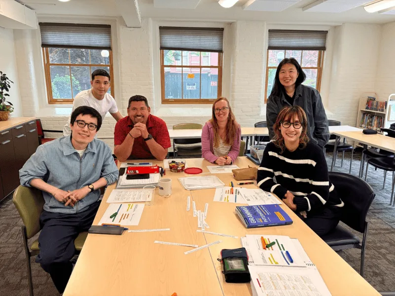 Six people sit around a table in a classroom with papers and supplies.