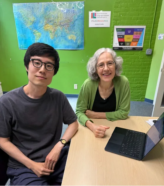 A young man in glasses and an older woman smile at a desk beside an open laptop, with maps and posters on a bright green classroom wall.